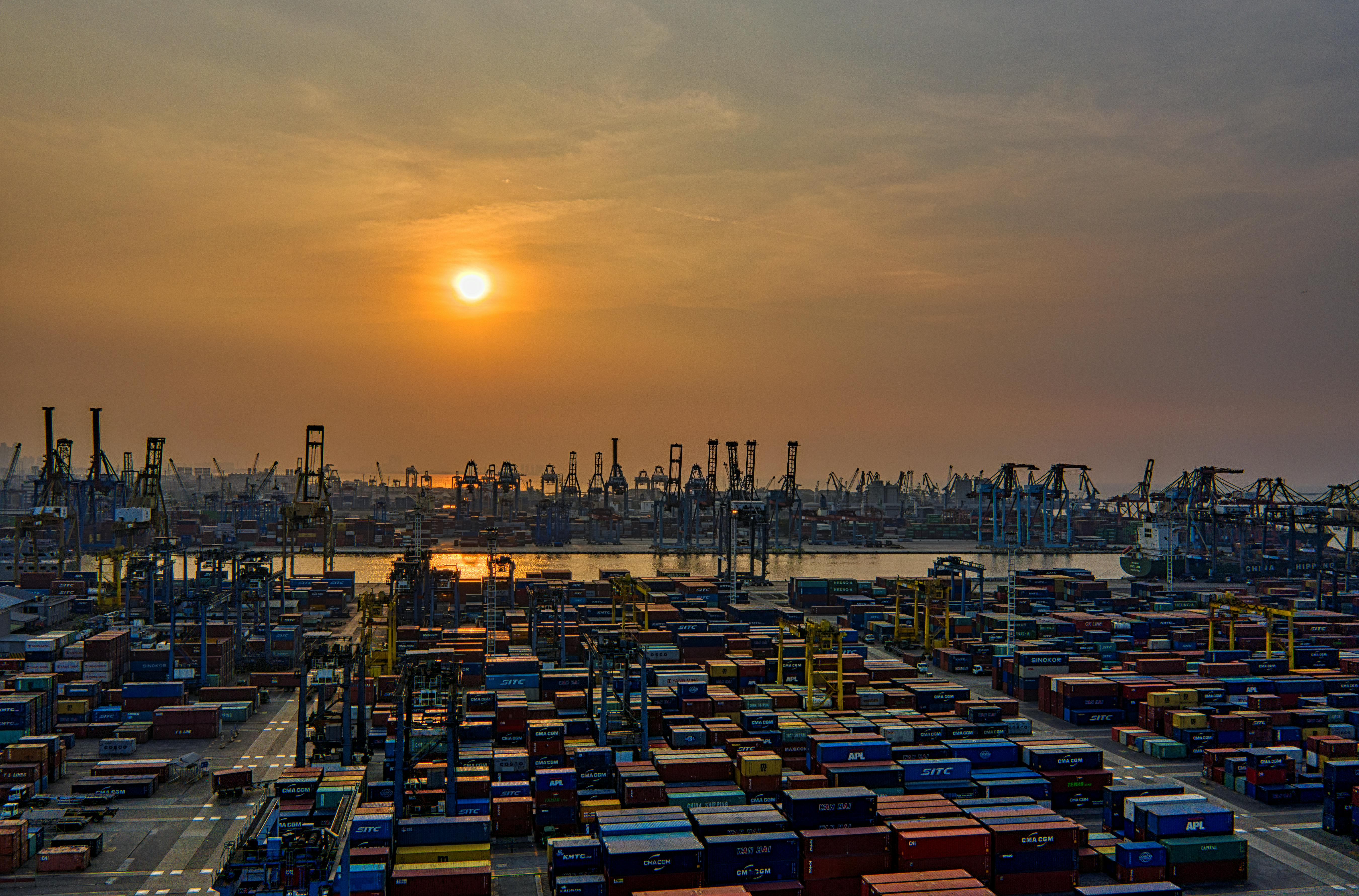pexels-photo-3057960-3057960 Sunset over a bustling seaport filled with stacked shipping containers and cranes.